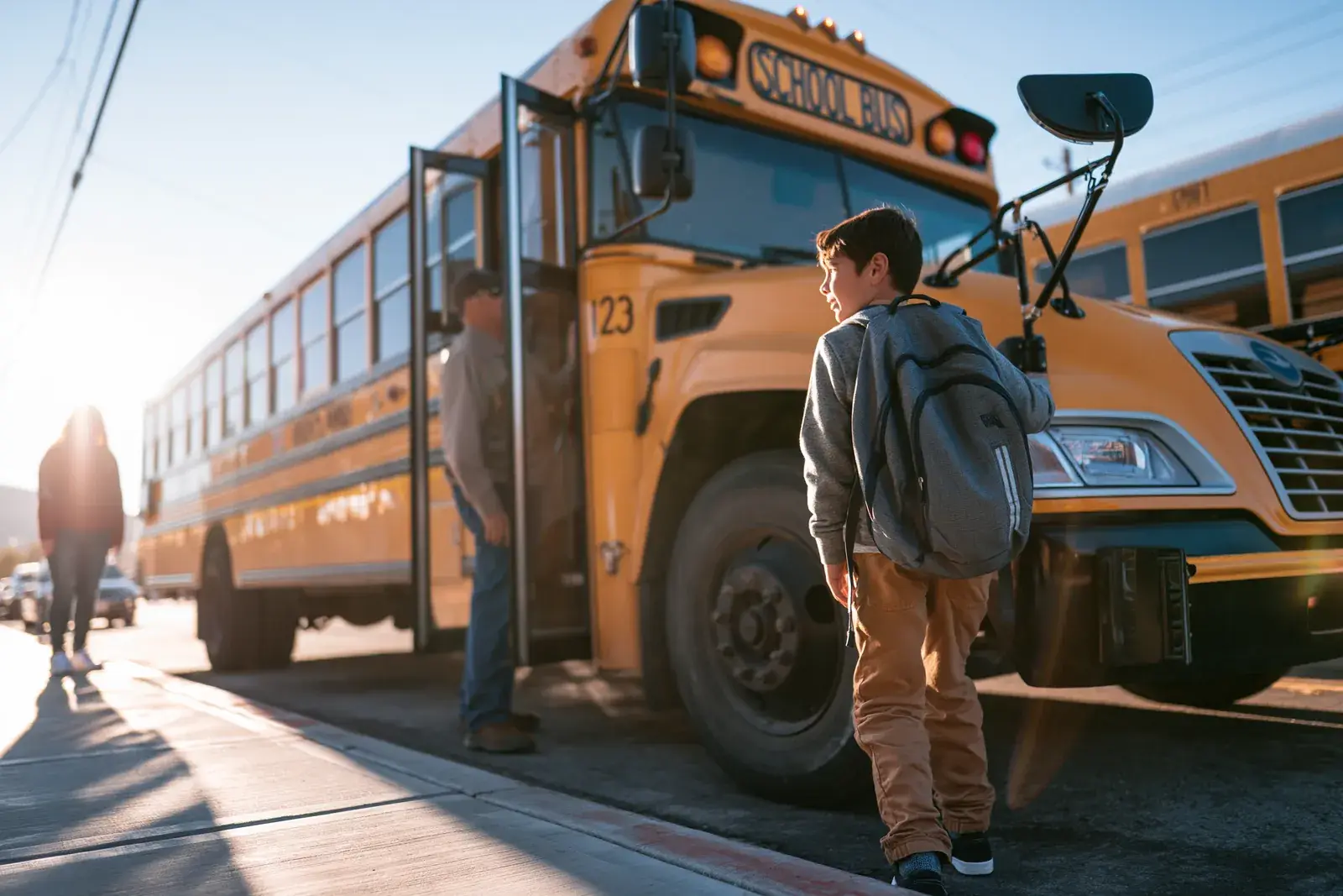 A boy with a backpack waiting at the open bus door, hard sun flare across the frame.